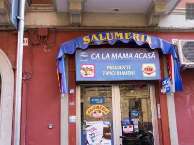 Il negozio si affaccia sulla strada. Sulla porta a vetri sono affissi i manifesti della compagnia telefonica rumena e l�insegna blu con la scritta in italiano �salumeria� e il nome �Ca la mama acasa � prodotti tipici rumeni�